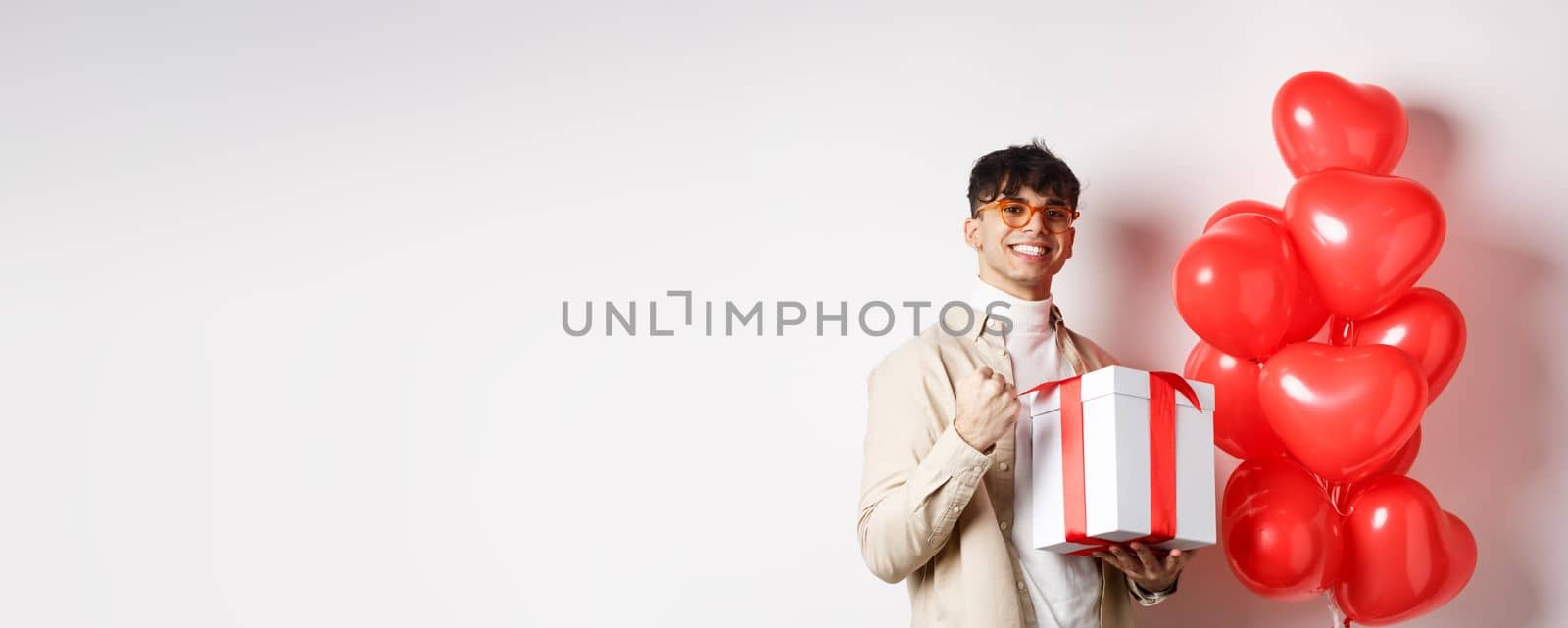 Valentines day and romance concept. Happy and confident boyfriend prepare gift for lover, saying yes and smiling, holding romantic gift, standing near red hearts balloons by Benzoix