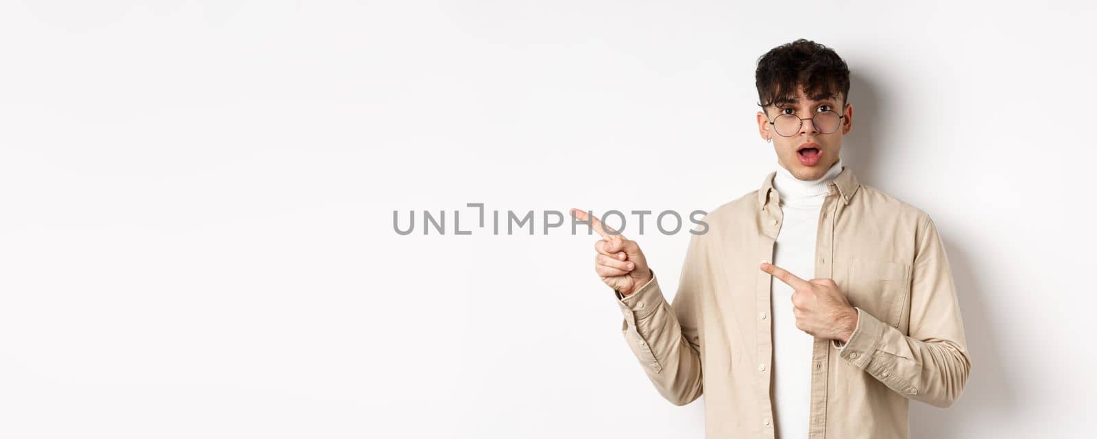 Portrait of young man in glasses drop jaw impressed, pointing fingers left at empty space with amazed face, standing on white background.