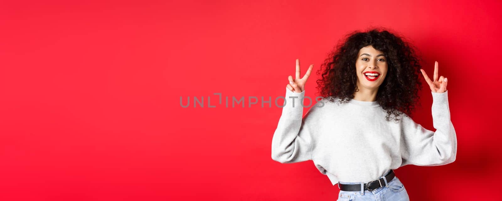 Stylish young woman with curly hairstyle, smiling happy and showing peace signs, standing in casual sweatshirt on red background.