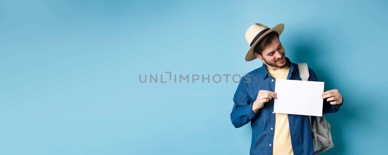 Happy tourist in straw hat smiling and looking at piece of paper for your logo, standing on blue background by Benzoix