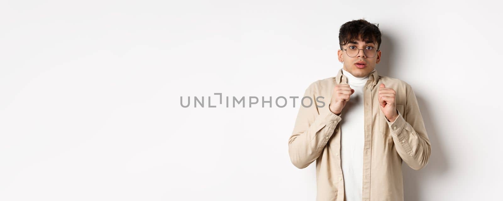 Startled man freeze from fear, gasping and looking scared at camera, standing in glasses on white background.