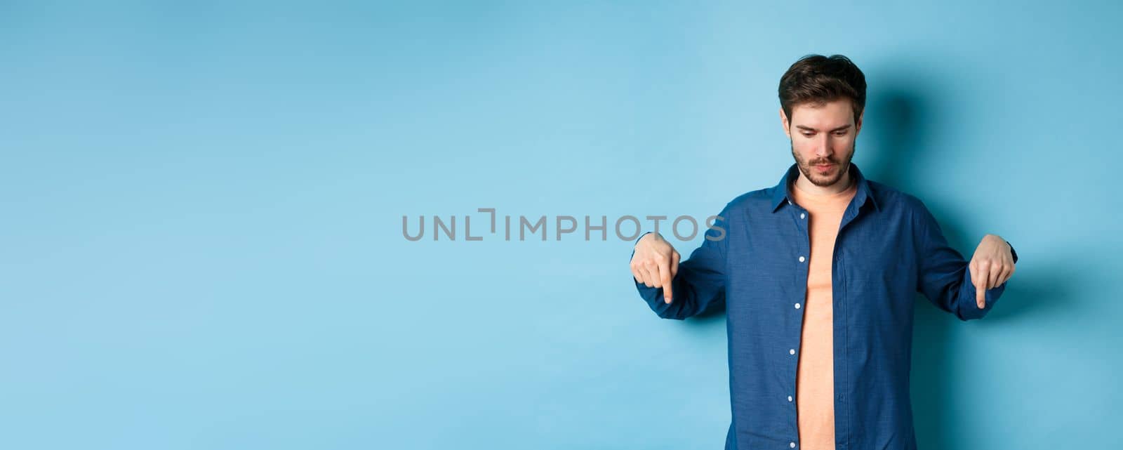 Image of modern guy in casual clothes, looking and pointing fingers down at empty space, reading logo, standing on blue background.