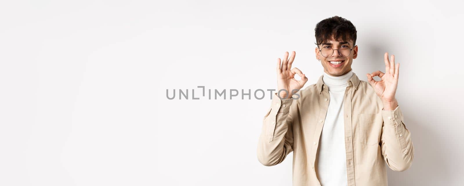 Handsome young man recommending good product, showing okay gestures and smiling, agree or approve something, praising excellent choice, standing on white background.