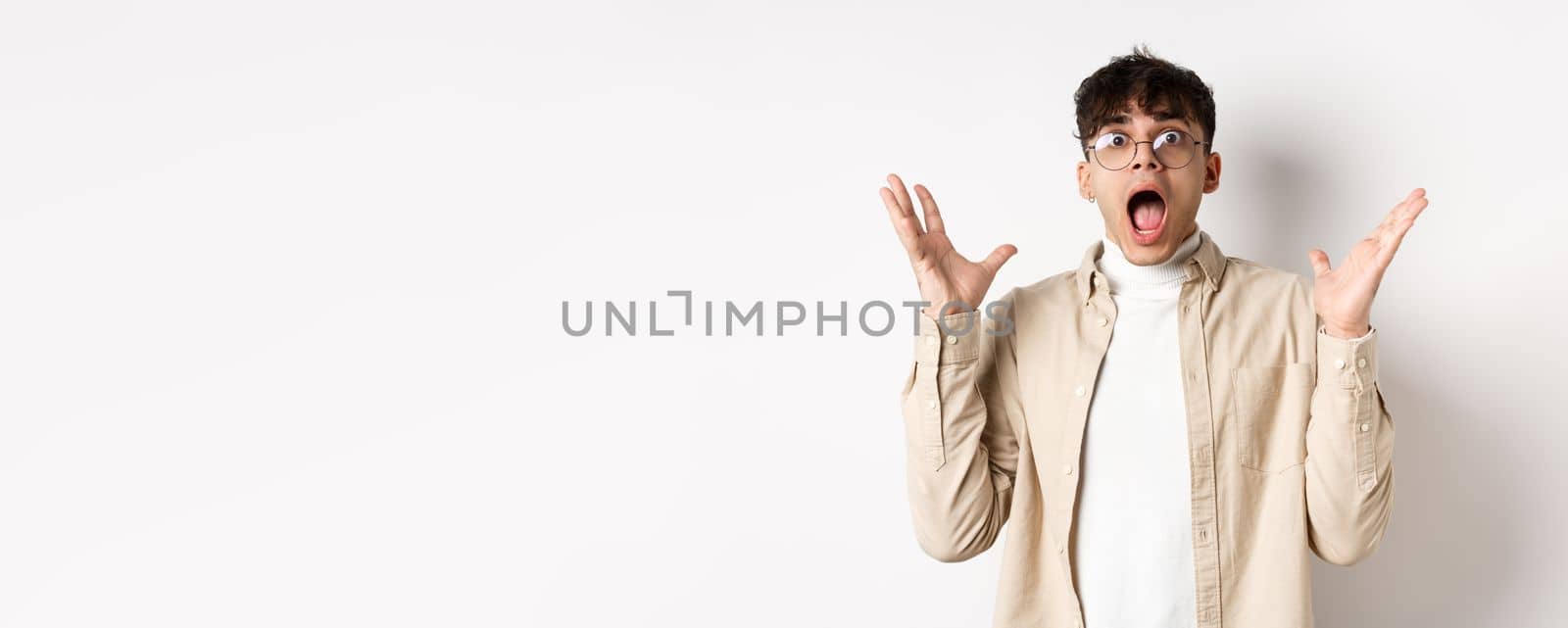 Scared young man in panic, screaming and looking axious, jumping startled and shocked, shaking hands nervously, standing on white background.