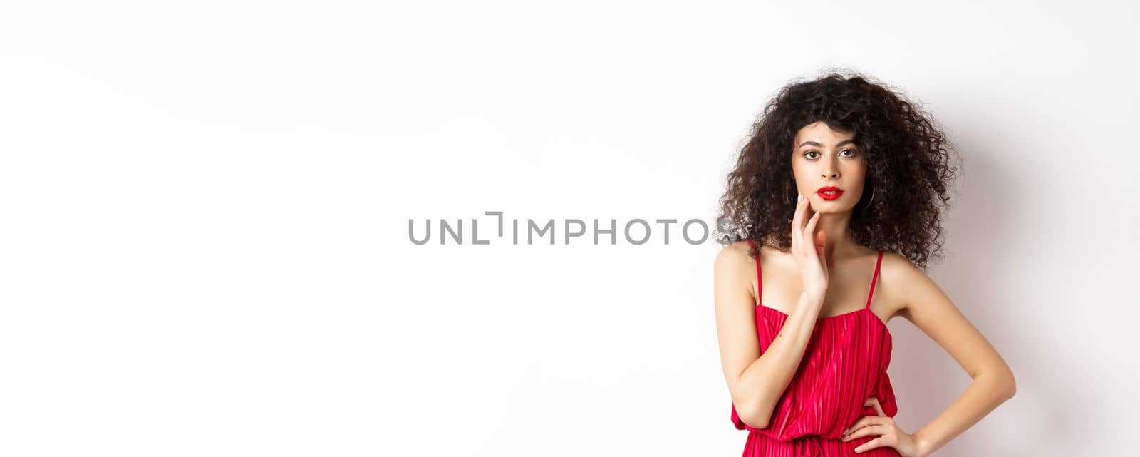 Beauty and fashion. Sensual female model with curly hair and red lips, wearing elegant dress, touching face and looking seductive, white background.
