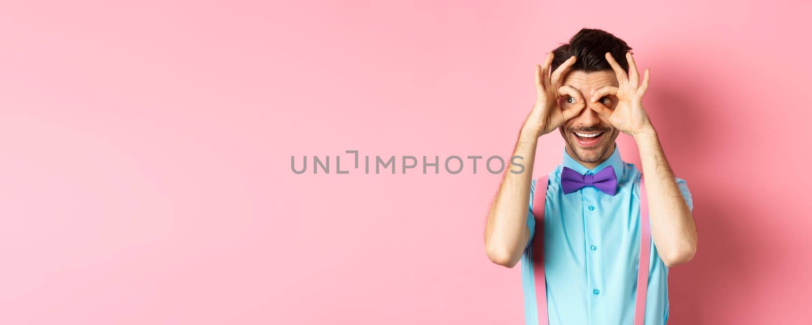 Cheerful man in bow-tie and suspenders looking through hand binoculars and smiling, seeing interesting holiday offer, staring at camera on pink background by Benzoix