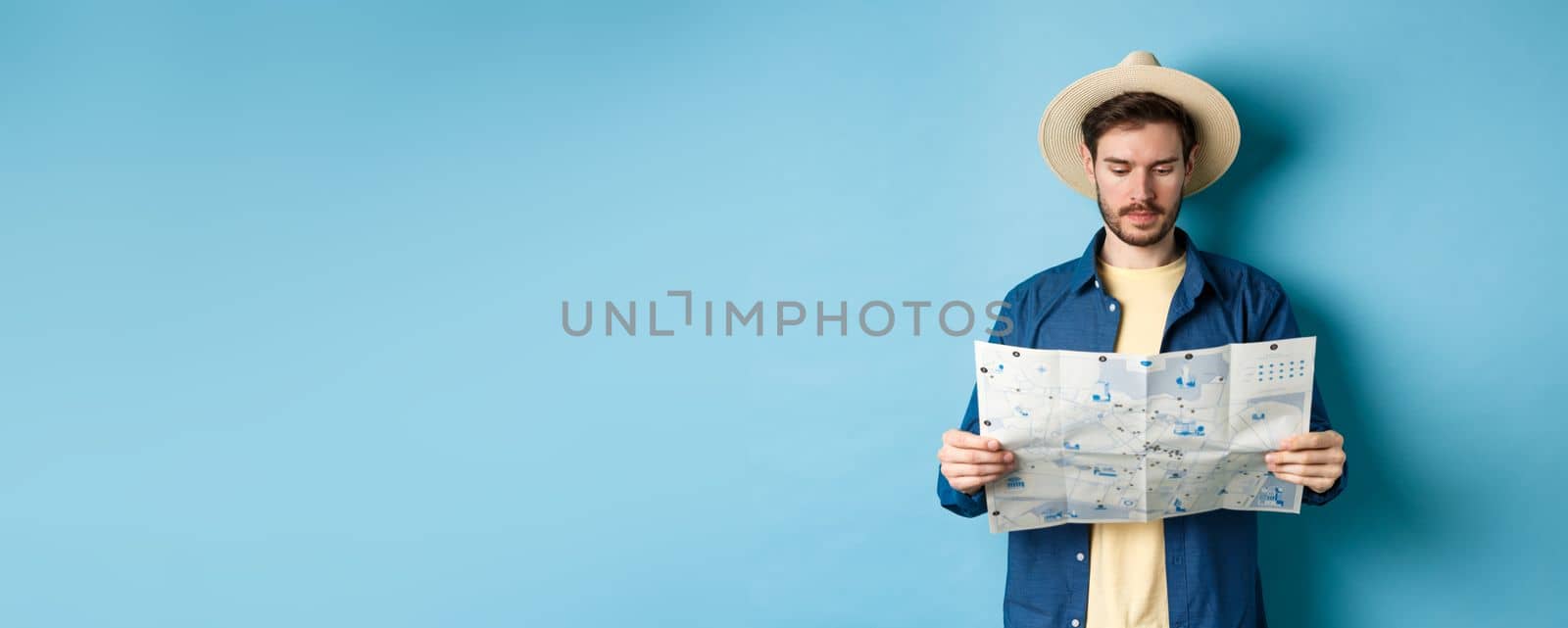 Handsome tourist with summer hat reading map, looking for sightseeing on vacation, standing on blue background.