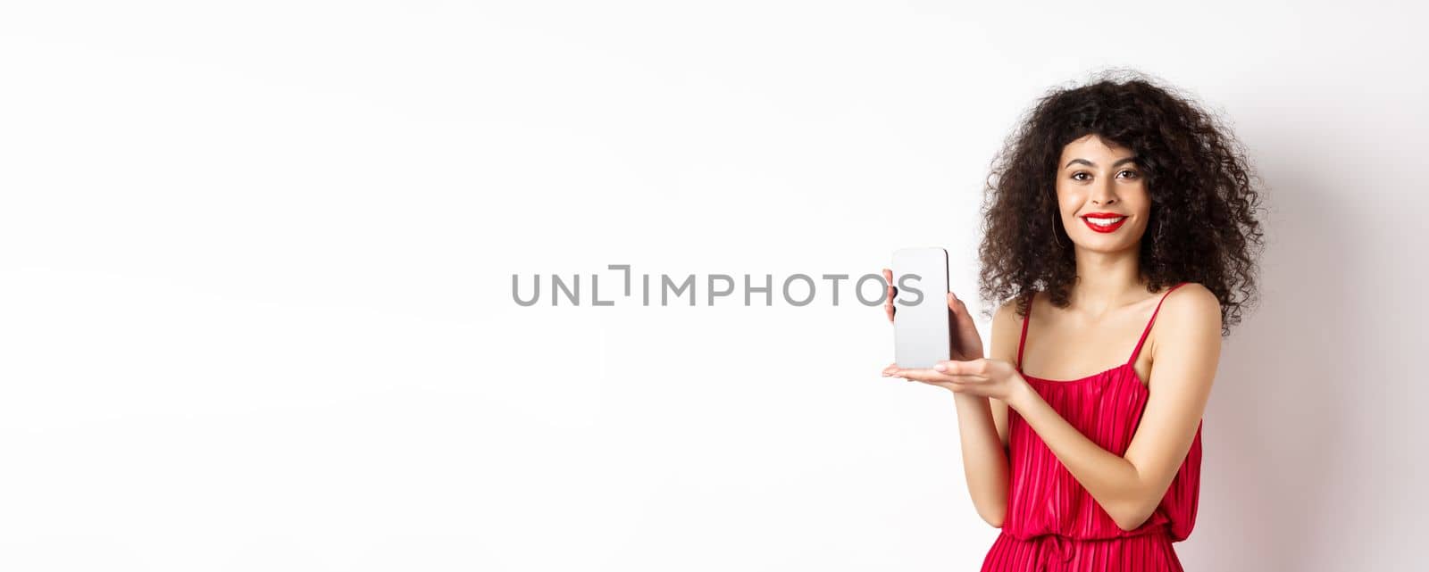 Elegant woman in red dress and makeup, showing blank smartphone screen and smiling, standing over white background by Benzoix