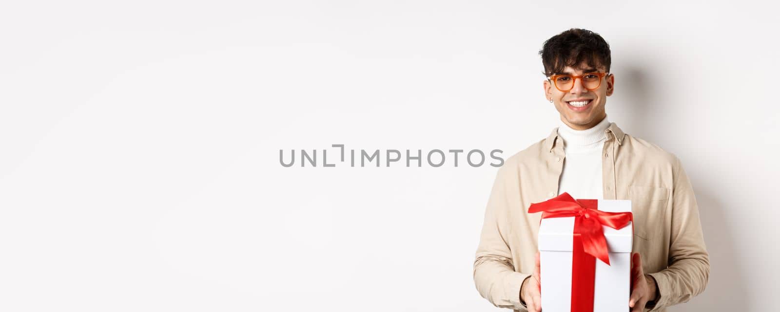 Handsome young man making a gift, holding box with present and smiling at camera, standing on white background.