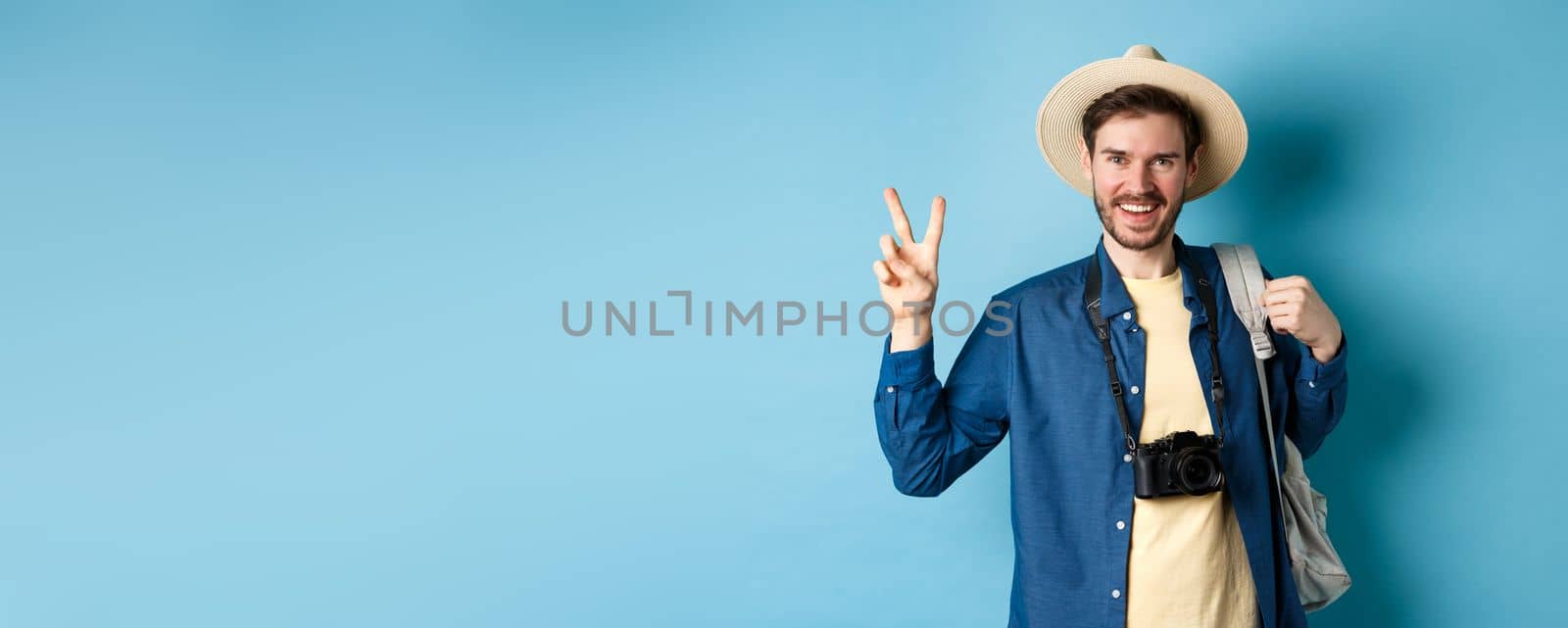 Happy handsome man taking photo on summer vacation, showing peace sign and smiling, wearing straw hat and holding tourist backpack, blue background by Benzoix