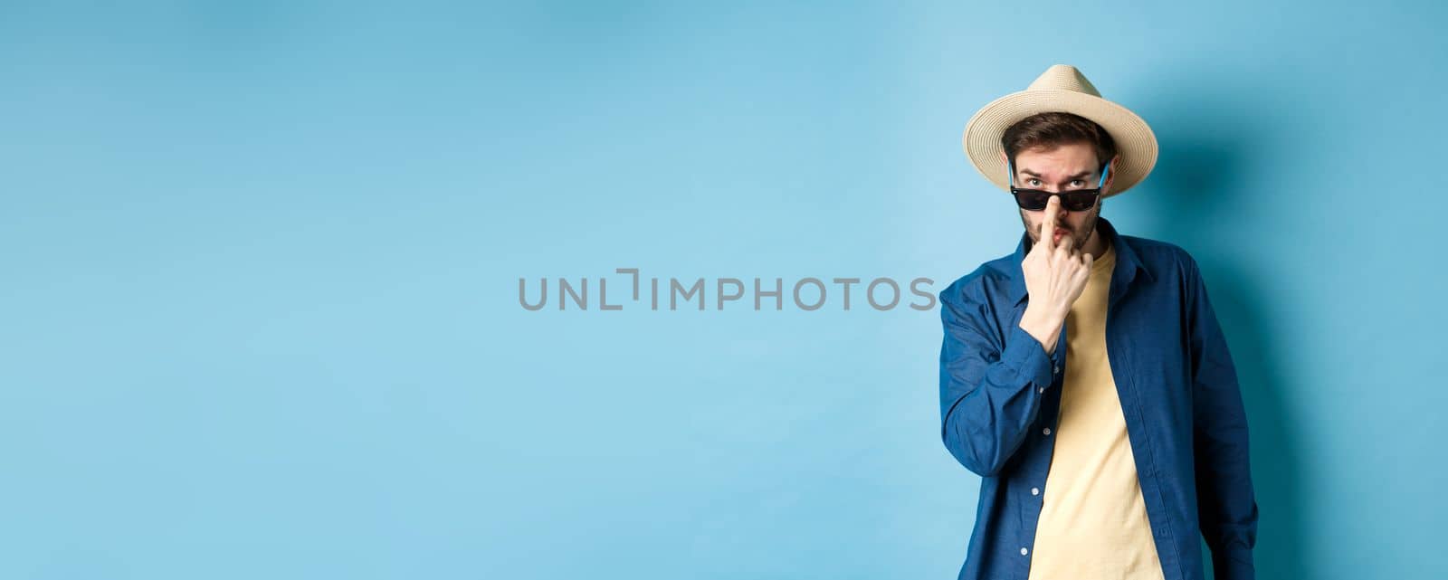 Handsome and cheeky guy put on sunglasses and straw hat, standing ready for summer vacation or tropical holiday, blue background.