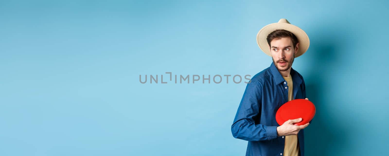 Handsome young guy in summer hat playing frisbee, throwing it aside, standing on blue background by Benzoix