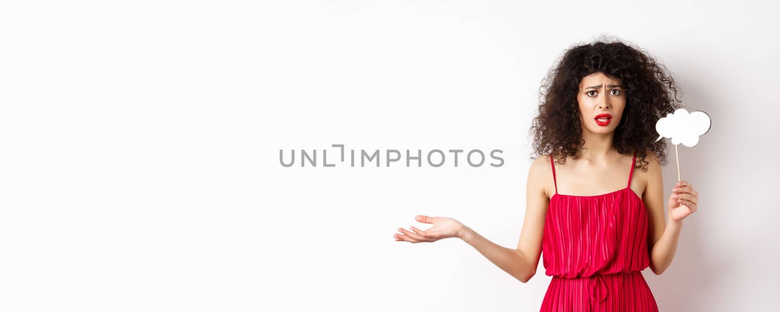 Confused young woman in red dress, frowning and looking upset, holding comment cloud stick, standing against white background by Benzoix