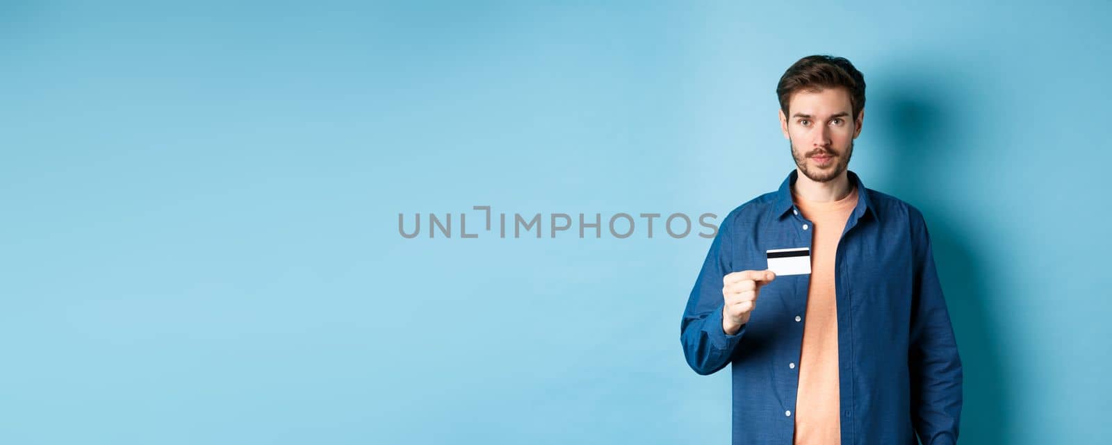 Handsome young man with beard, wearing casual clothes, showing plastic credit card and looking at camera, blue background.