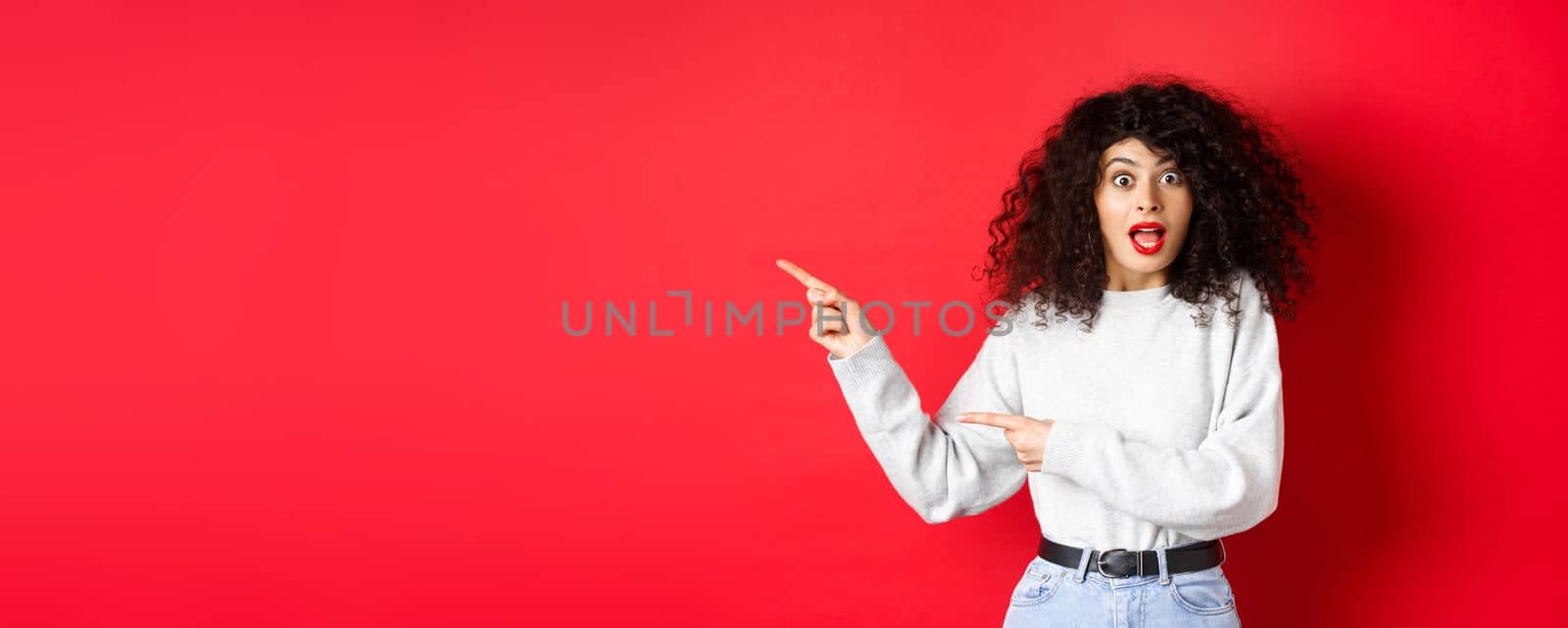 Excited caucasian woman in sweatshirt, checking out banner, pointing fingers left at empty space, showing advertisement, red background.