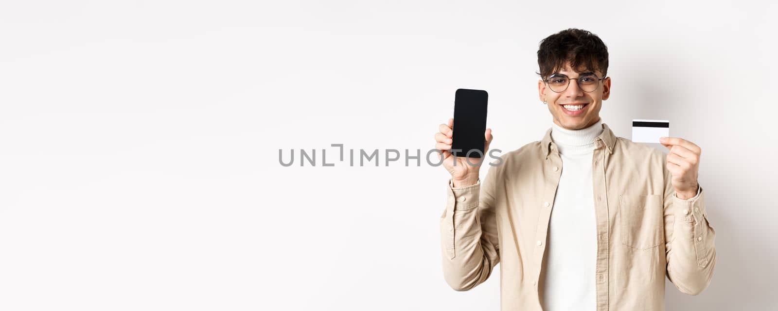 Online shopping. Natural guy in glasses showing empty smartphone screen and plastic credit card, smiling pleased, recommending bank, standing on white background.