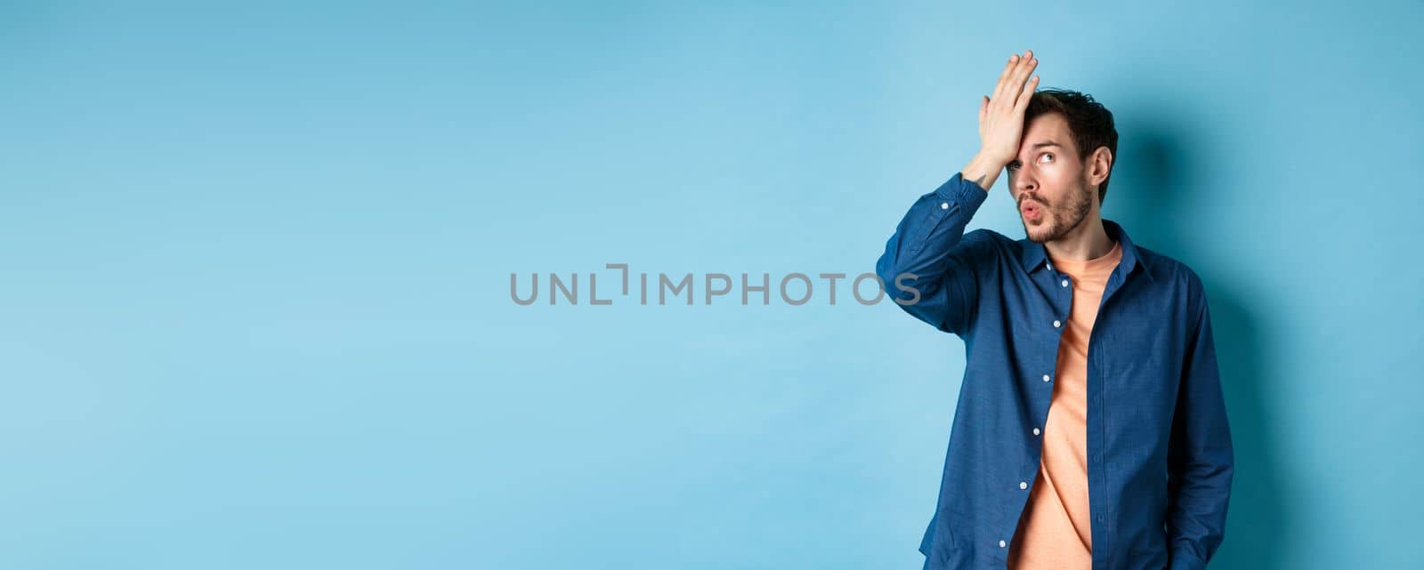 Annoyed man making facepalm and roll eyes from something stupid, standing over blue background with hand on forehead.