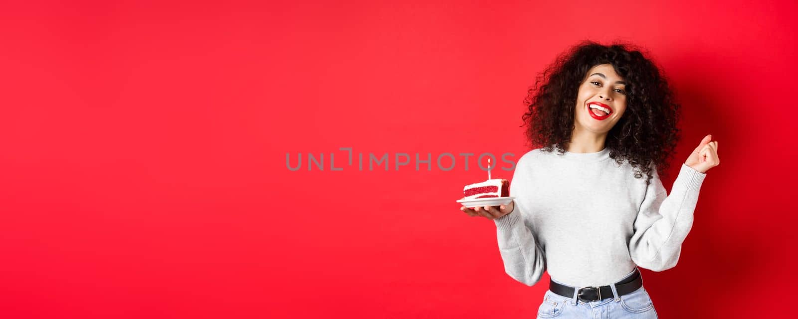 Celebration and holidays concept. Happy beautiful woman dancing and making birthday wish, holding b-day cake and smiling, standing on red background.