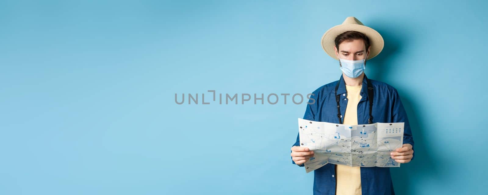 Covid-19, pandemic and travel concept. Young man in medical mask reading tourist map, searching for route to sightseeings, standing on blue background by Benzoix