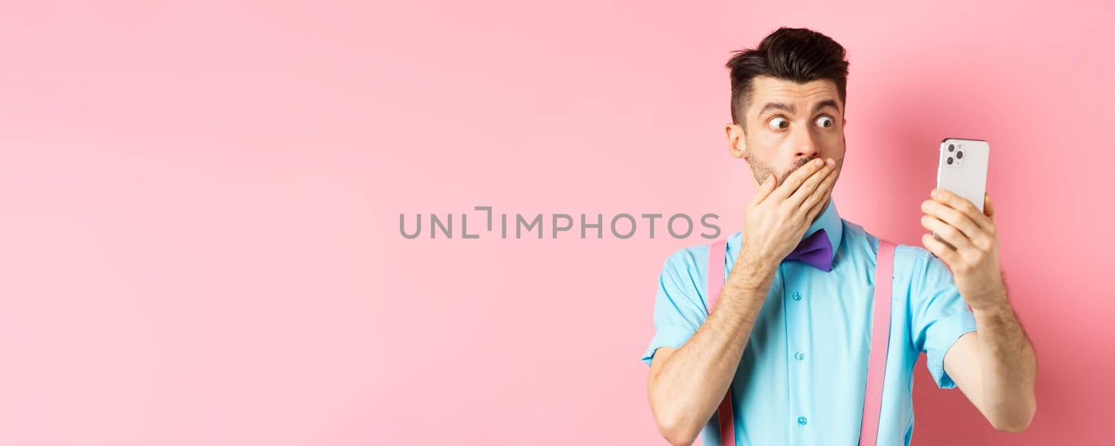 Technology concept. Silly young man looking shocked at smartphone screen while having video chat, standing on pink background and cover mouth with hand.