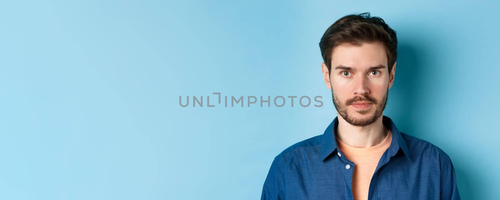 Close-up of young man with beard looking serious at camera, standing in casual shirt on blue background.