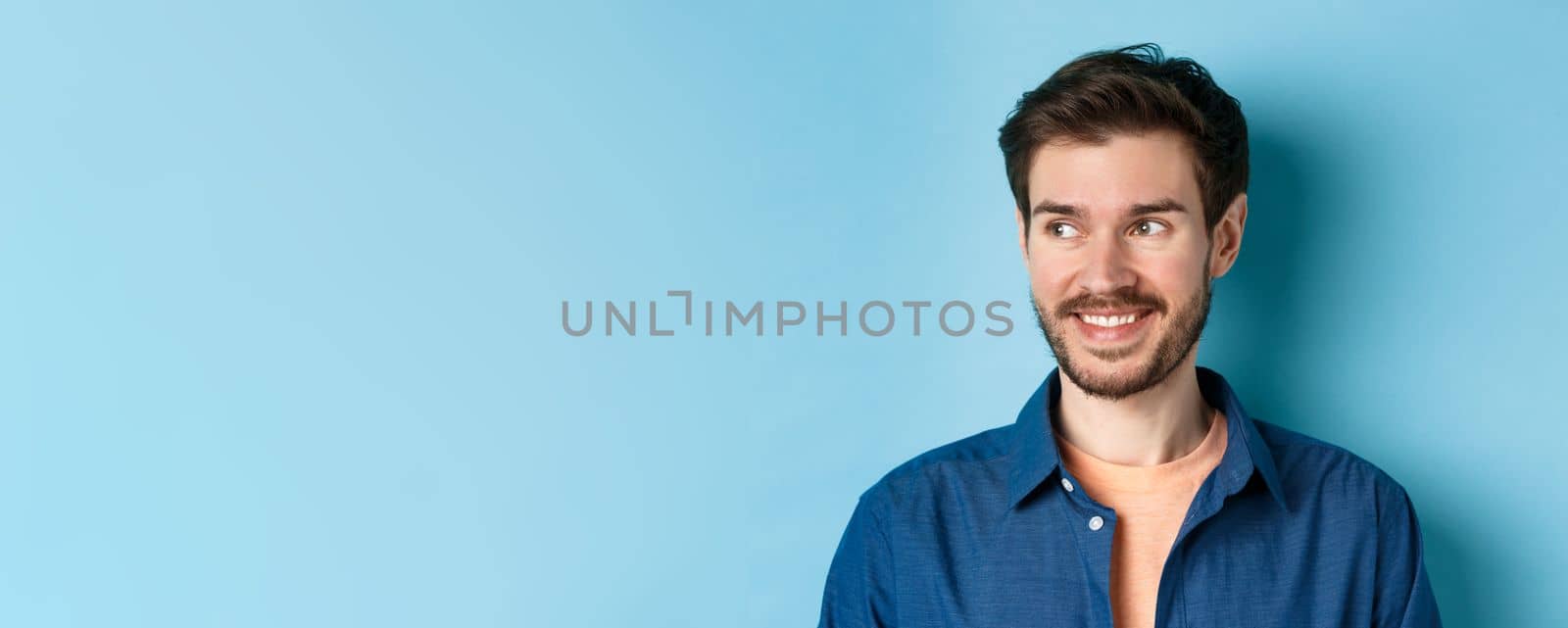 Close up of handsome confident man with beard, looking at empty space and smiling, standing on blue background.