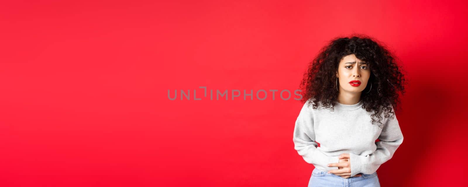 Image of young woman having stomach ache, bending from pain and complaining on painful menstrual cramps, standing on red background by Benzoix