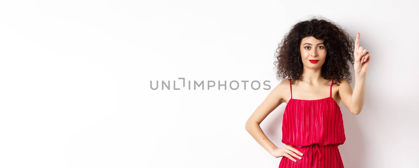 Smiling caucasian woman with curly hair, wearing red dress, showing rule number one gesture, raising finger and looking confident at camera, white background by Benzoix
