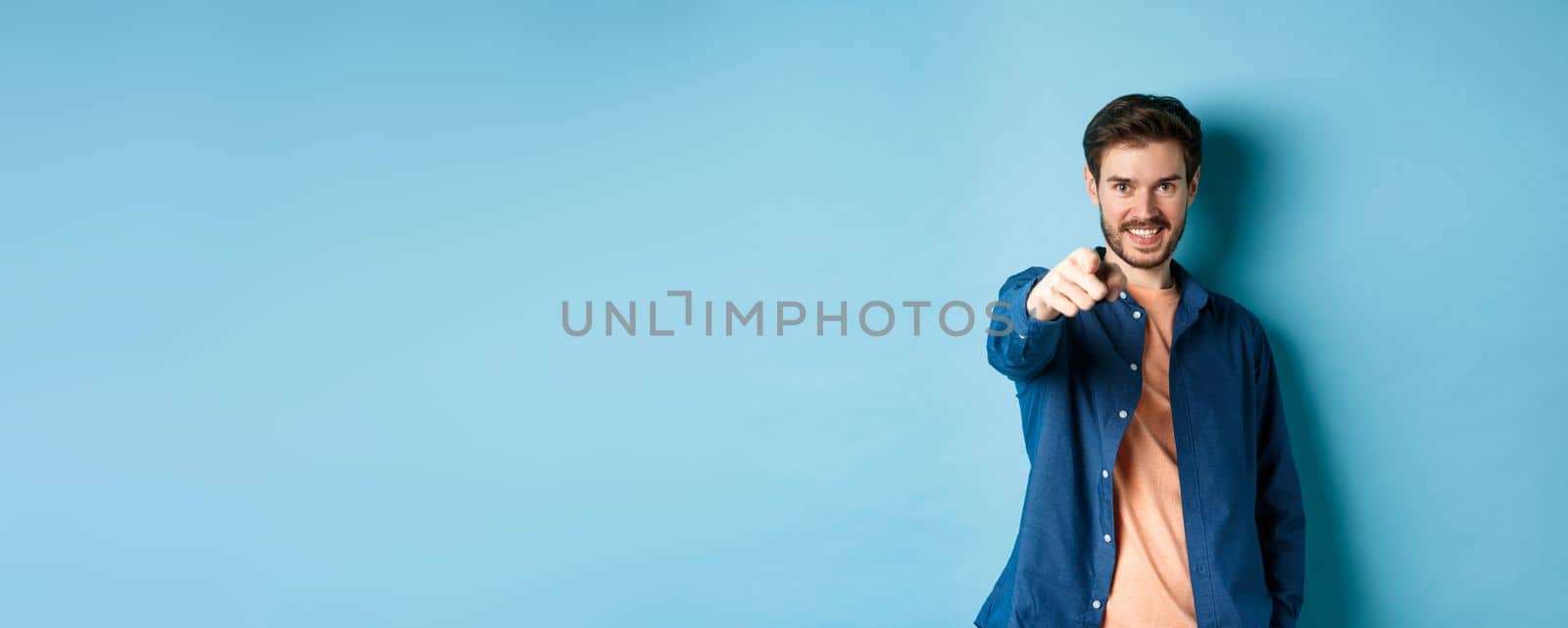 Confident smiling guy choosing or inviting you, pointing finger at camera decisive, standing on blue background.