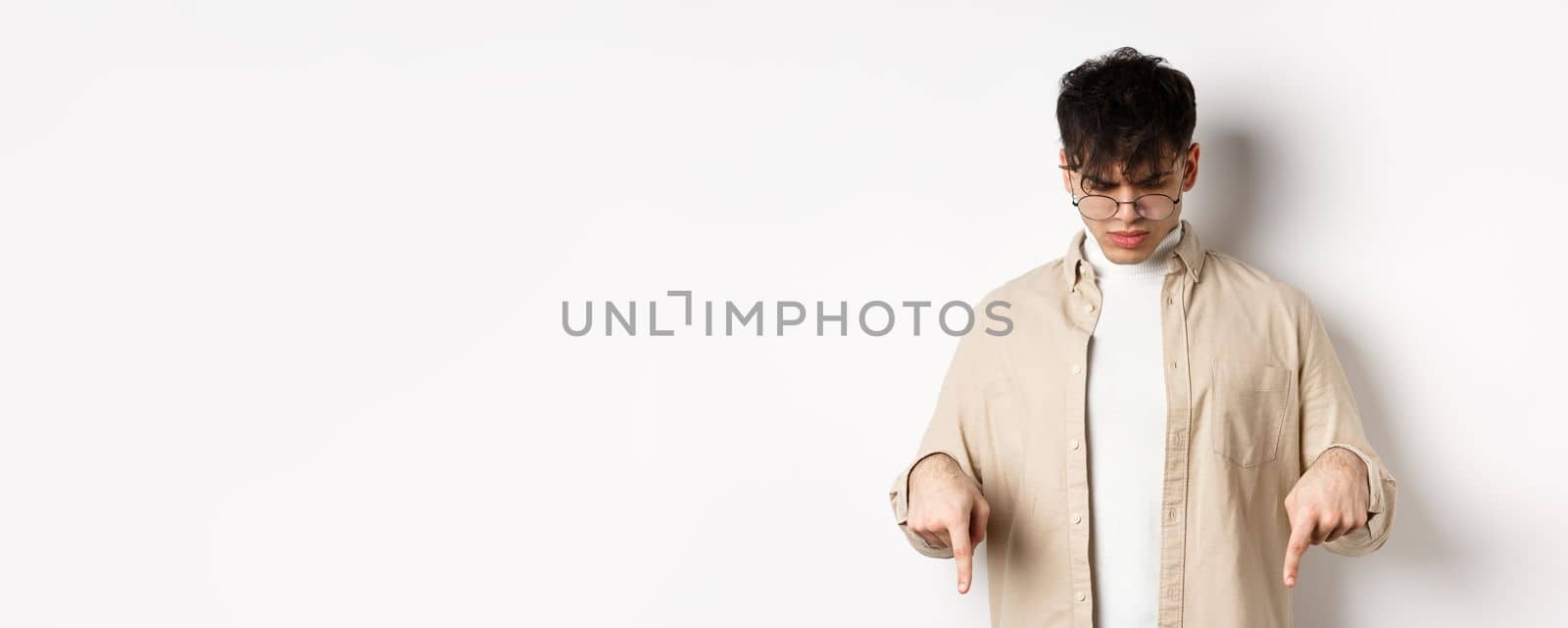 Confused frowning guy in glasses looking, pointing fingers down at something strange, standing puzzled on white background by Benzoix