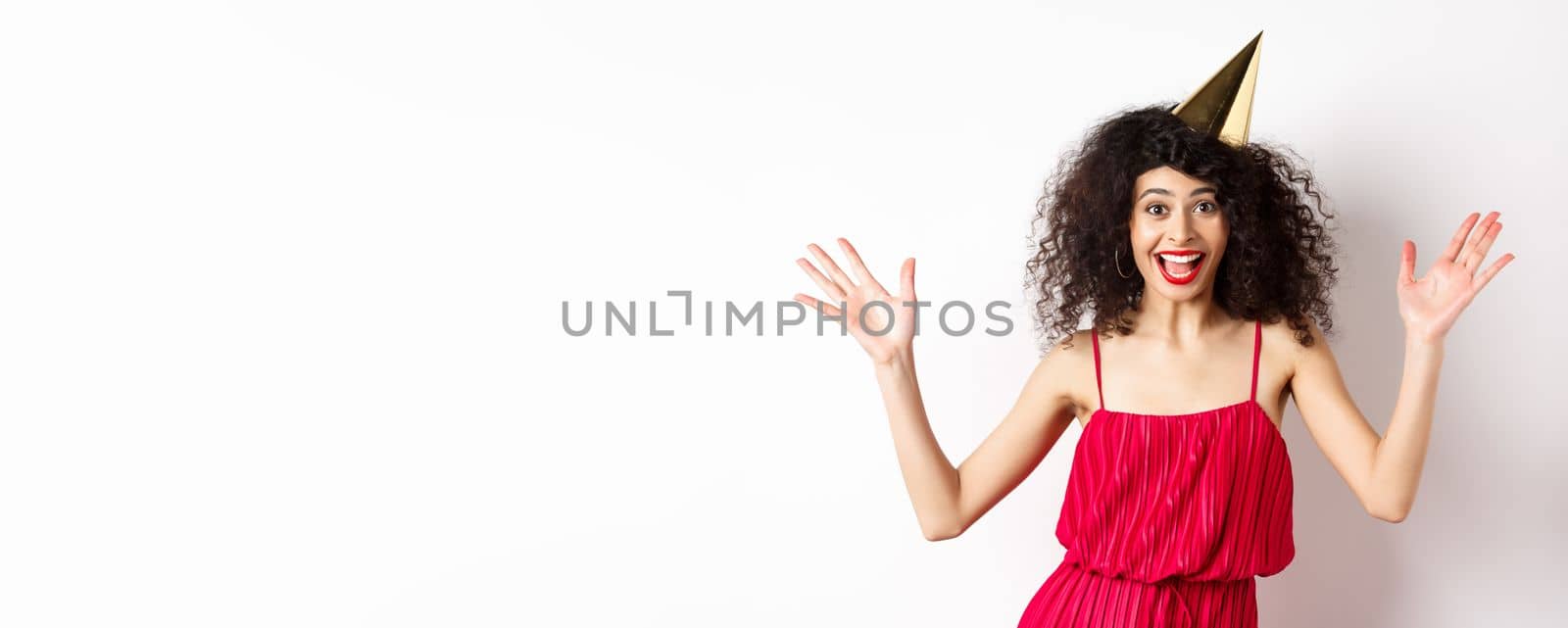 Cheerful young woman in red dress, celebrating birthday, wearing party hat and smiling, screaming of joy, standing on white background.