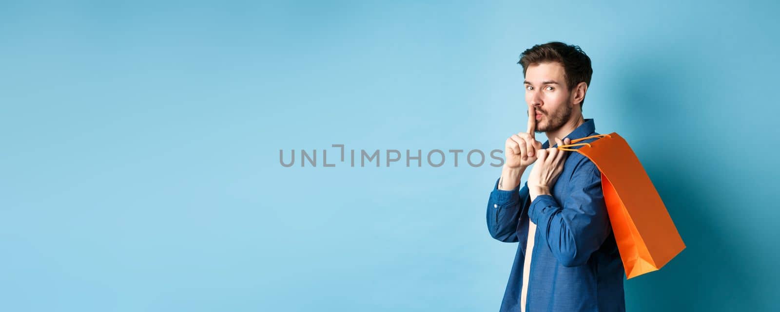 Image of modern guy holding shopping bag on shoulder, turn behind and shushing at camera, telling a secret, making surprise, standing on blue background.
