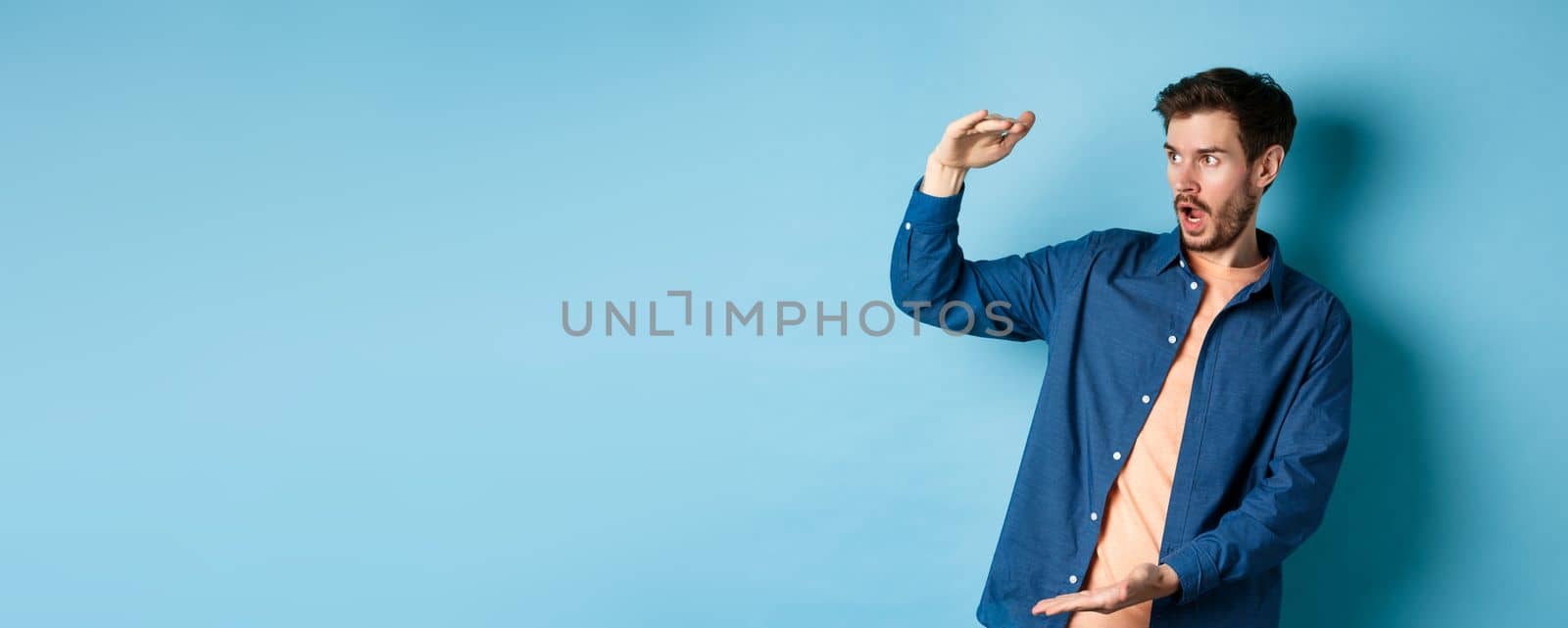 Impressed caucasian guy staring at empty space with big object, shaping large size, standing on blue background by Benzoix