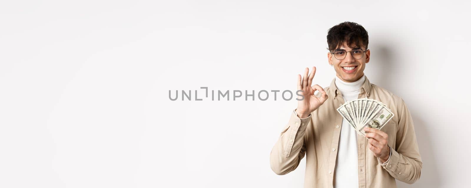 Handsome modern man in glasses showing dollar bills and okay gesture, making money, standing with cash on white background by Benzoix