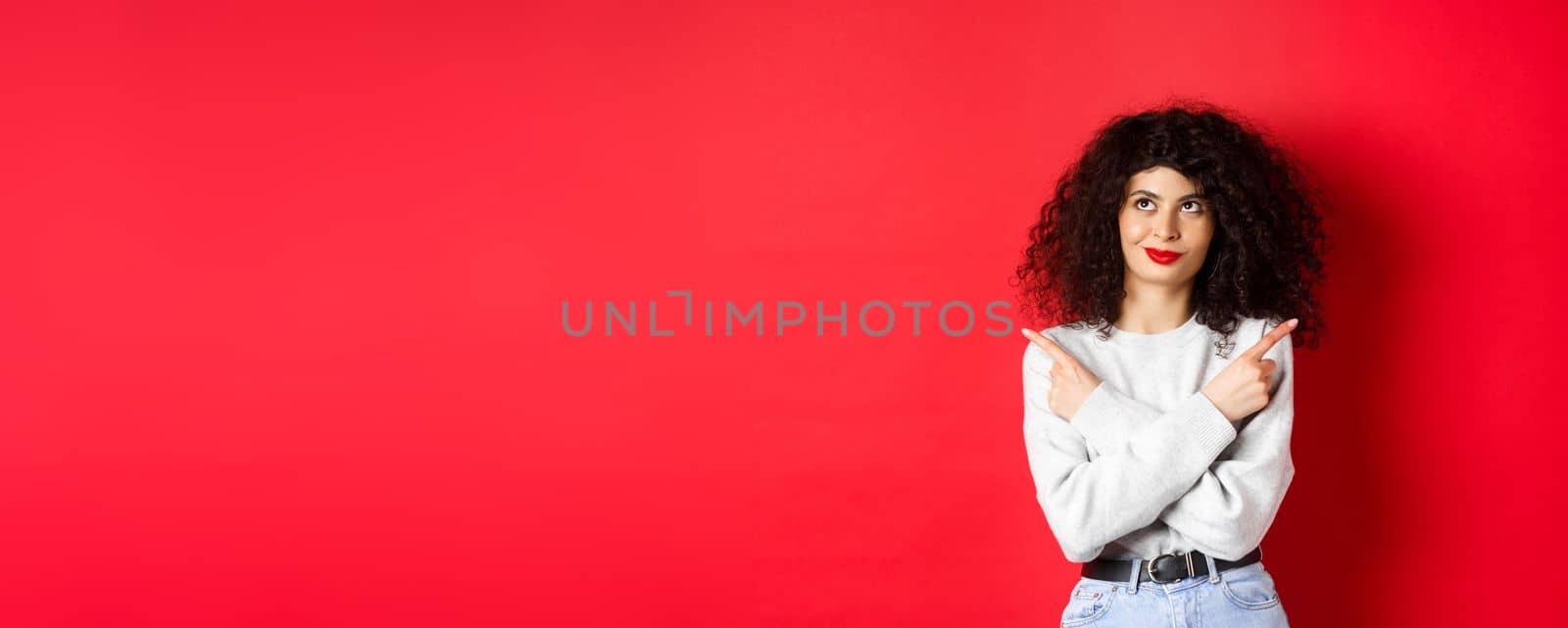 Pensive young woman making choice, pointing fingers sideways and looking at upper left corner with confident face, standing on red background by Benzoix