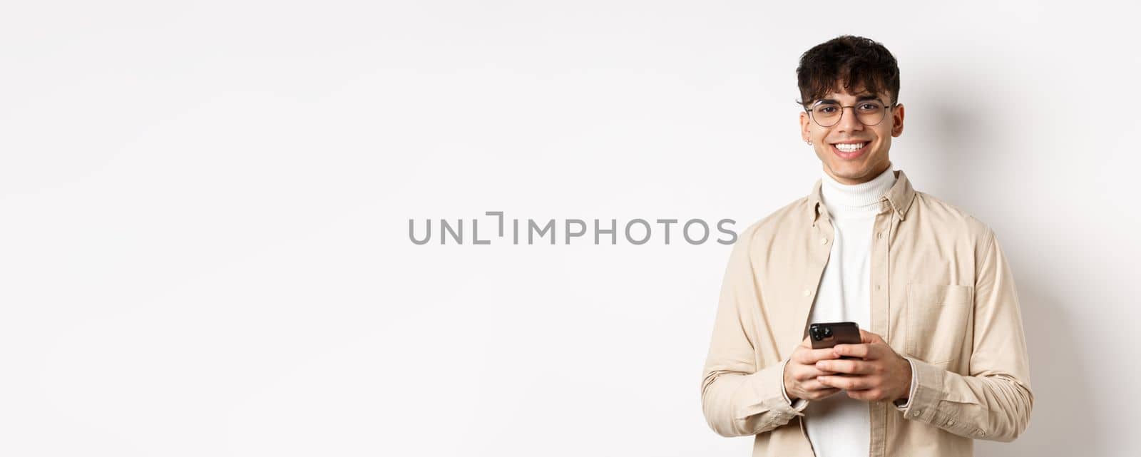 Real people. Handsome guy in glasses using mobile phone and smiling happy at camera, standing on white background by Benzoix
