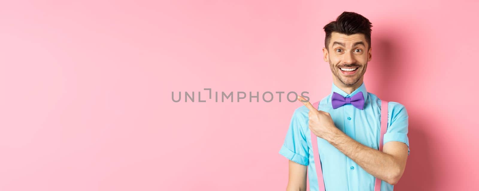 Portrait of handsome happy man showing logo, pointing left and smiling at camera, recommending shop offer, standing on pink background.