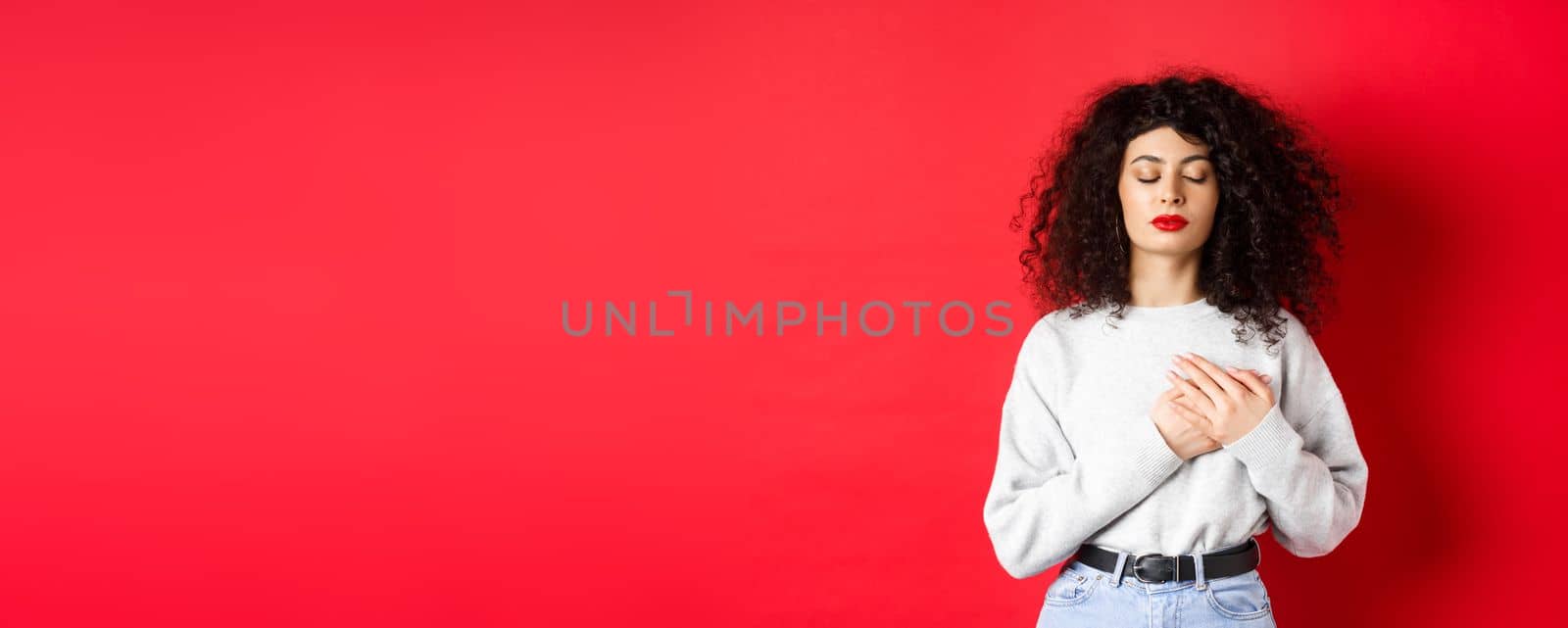 Image of calm young woman with curly hairsty, close eyes and holding hands on heart, keeping warm memories, feeling nostalgic, standing on red background.