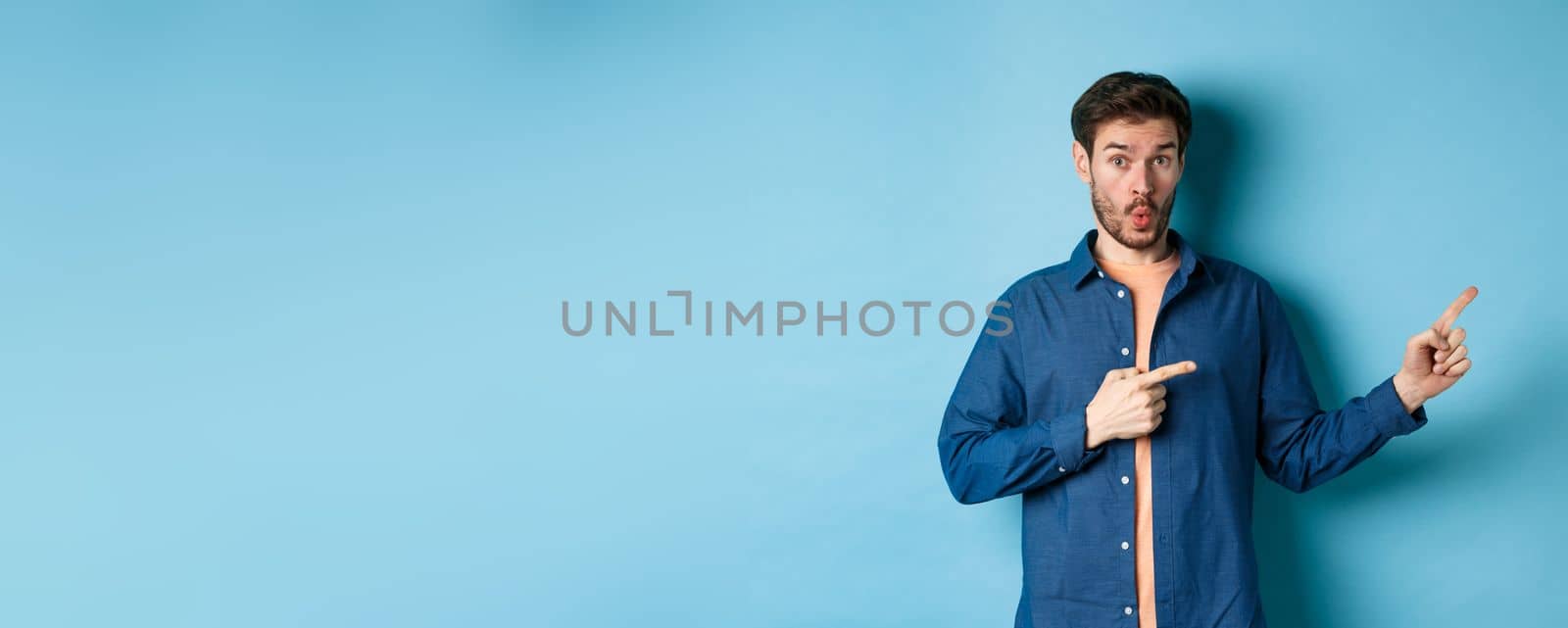 Surprised young man checking out promo, pointing fingers right at logo and saying wow, standing on blue background by Benzoix