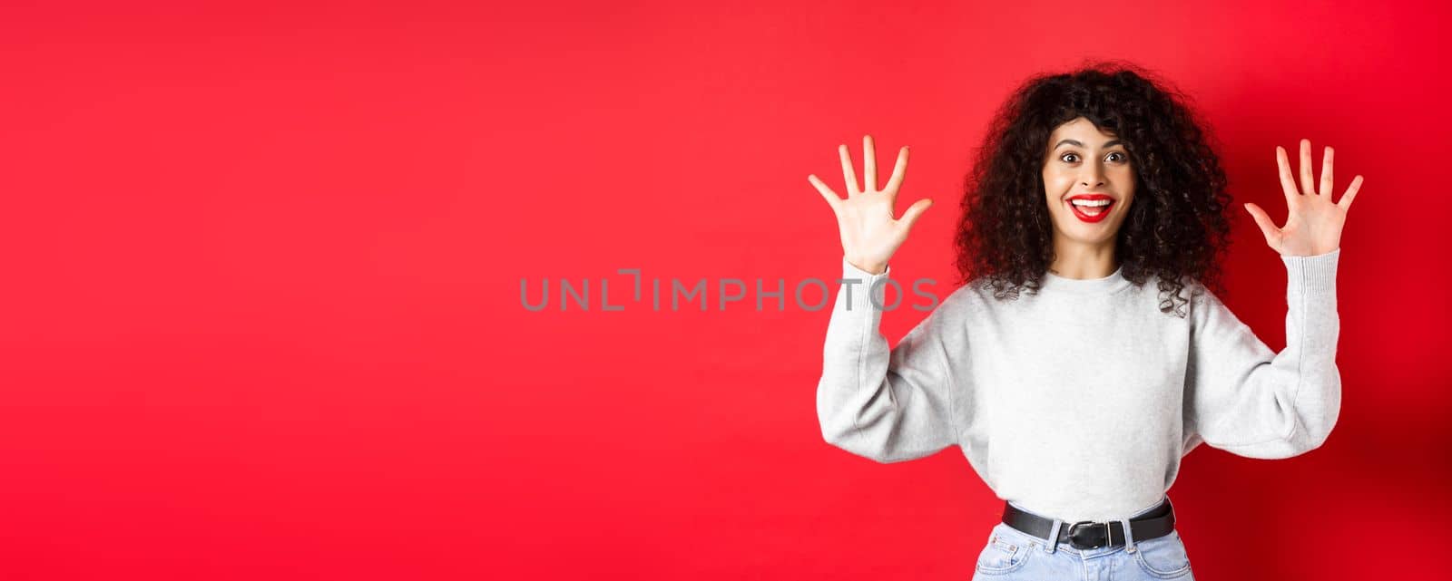 Happy beautiful woman smiling and raising hands up, showing number ten, ordern dozen of something, standing in sweatshirt on red background by Benzoix