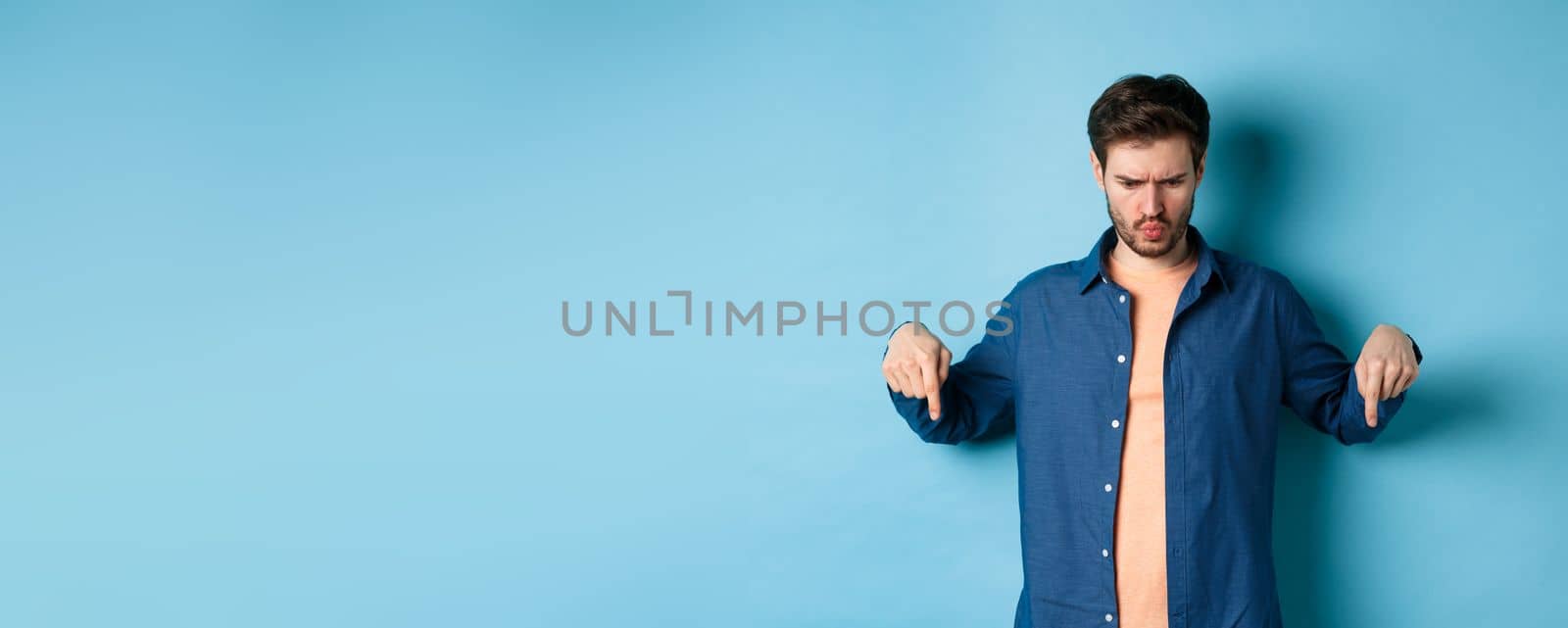 Confused and shocked guy looking and pointing down at something strange, standing on blue background. Copy space