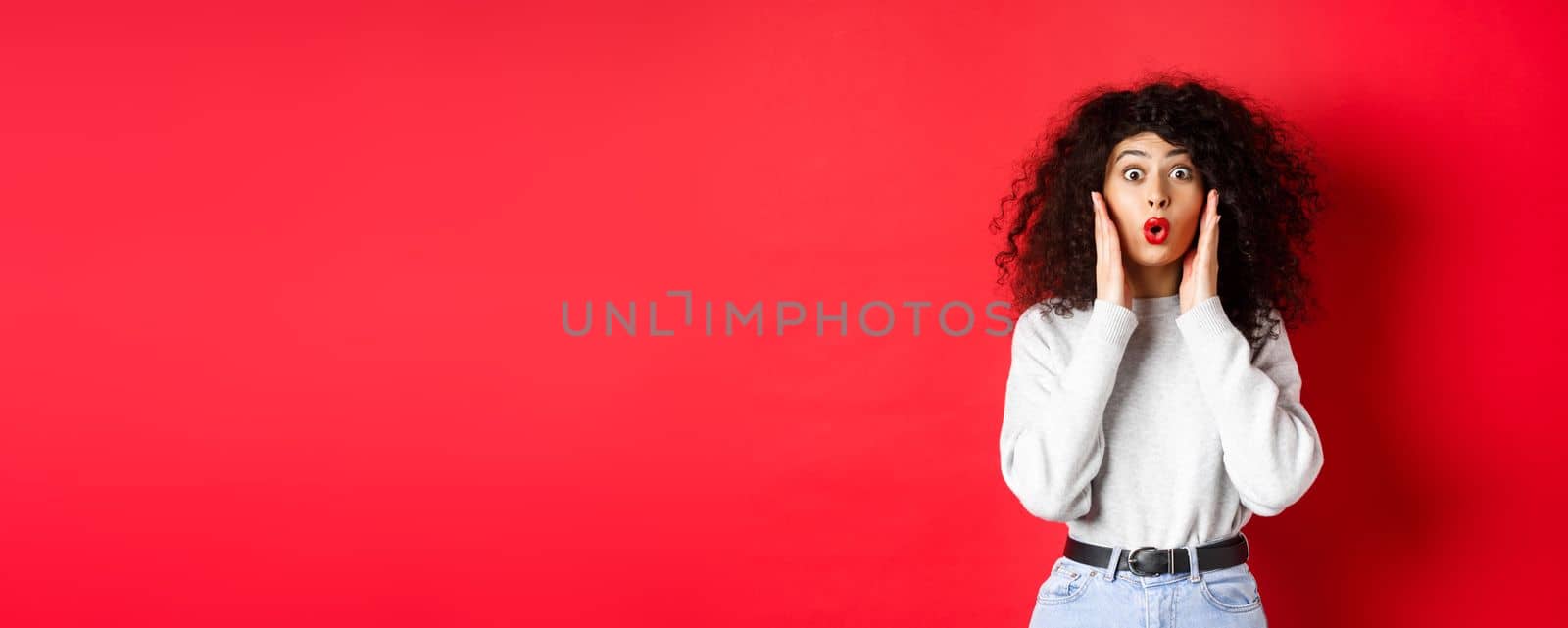 Surprised young woman hear amazing news, looking at promo and saying wow, stare with disbelief, standing on red background by Benzoix