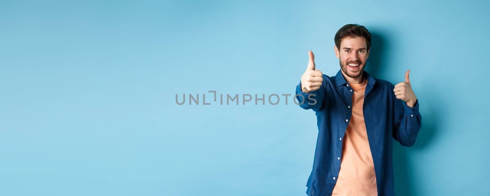 Positive smiling guy showing thumbs up and smiling, complimenting you, praising good job, well done gesture, standing on blue background.