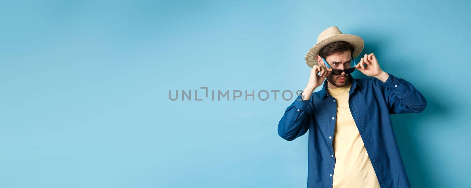 Portrait of guy on vacation staring aside with shocked and cringe face, take-off sunglasses and look at empty space on blue background by Benzoix