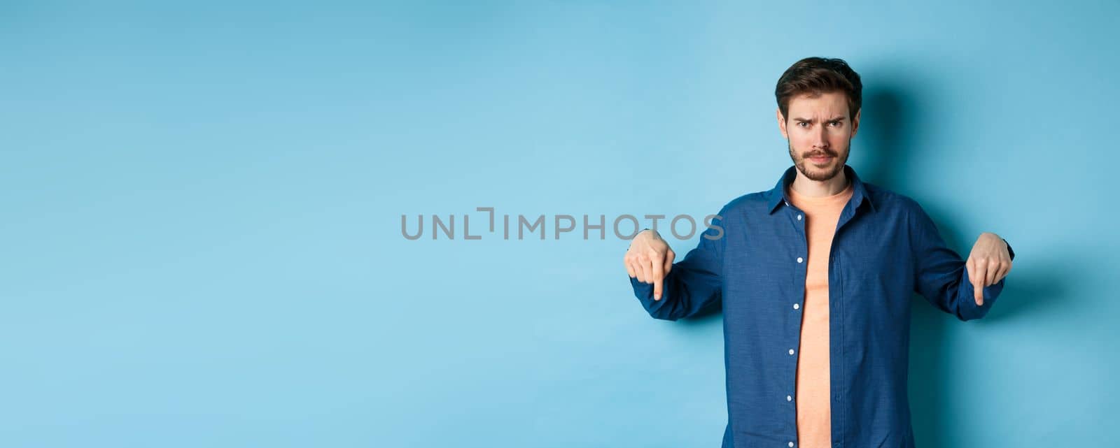 Concerned young man, frowning and looking worried, pointing fingers down, showing something bad, standing on blue background.