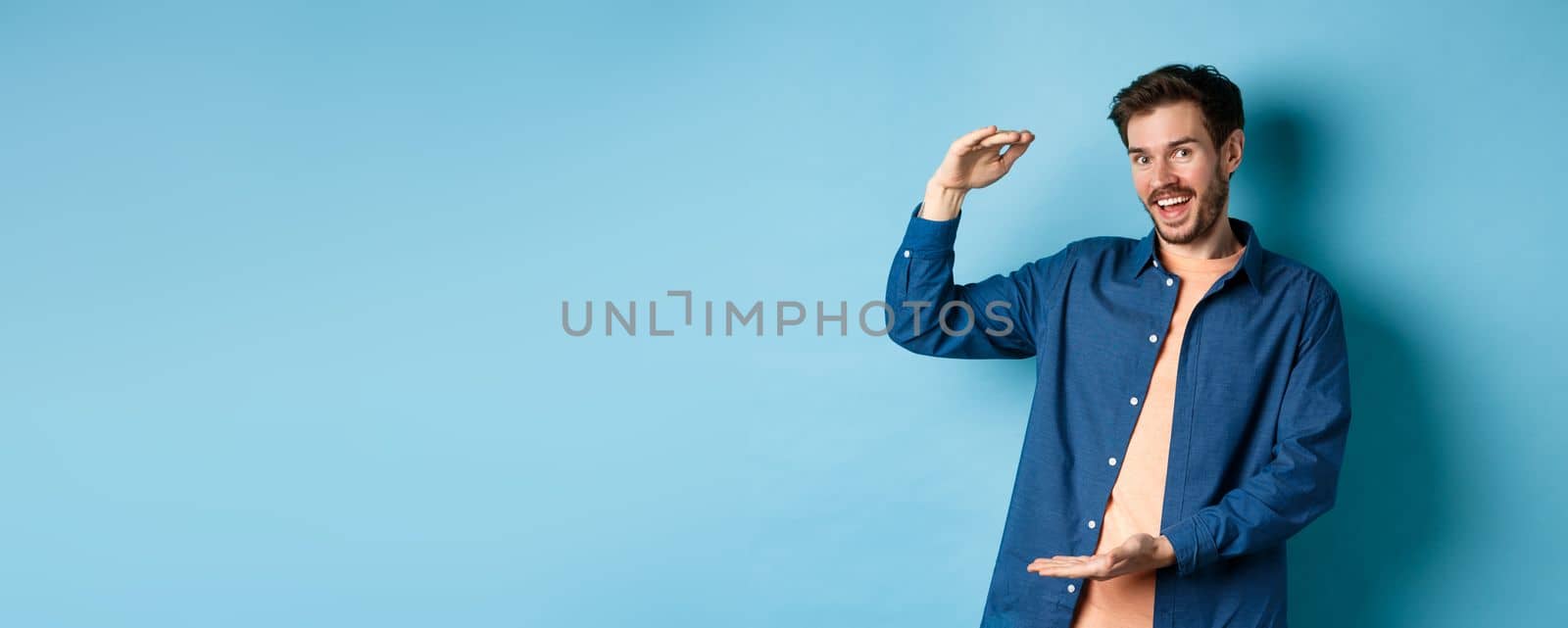 Cheerful modern guy smiling, showing big size and looking happy, standing on blue background. Copy space