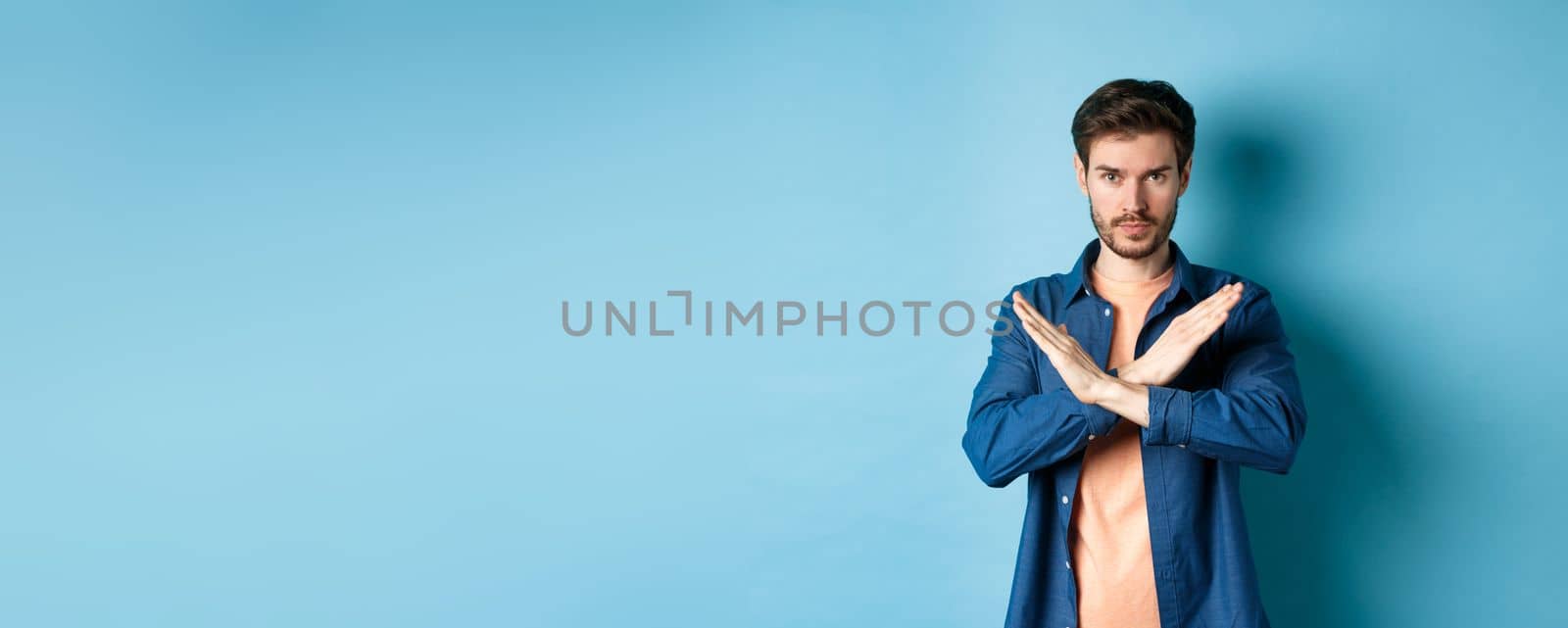 Serious caucasian man tell no, showing cross stop gesture and looking confident at camera, prohibit something bad, standing on blue background.