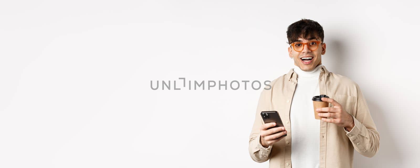 Cheerful smiling young man in glasses drinking coffee and holding smartphone, looking excited at camera, standing on white background.