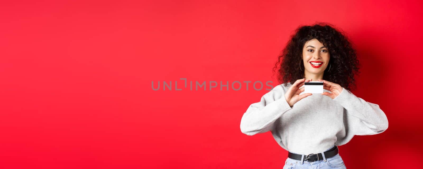 Happy lady with curly hair showing plastic credit card and smiling, standing against red background. Shopping concept by Benzoix