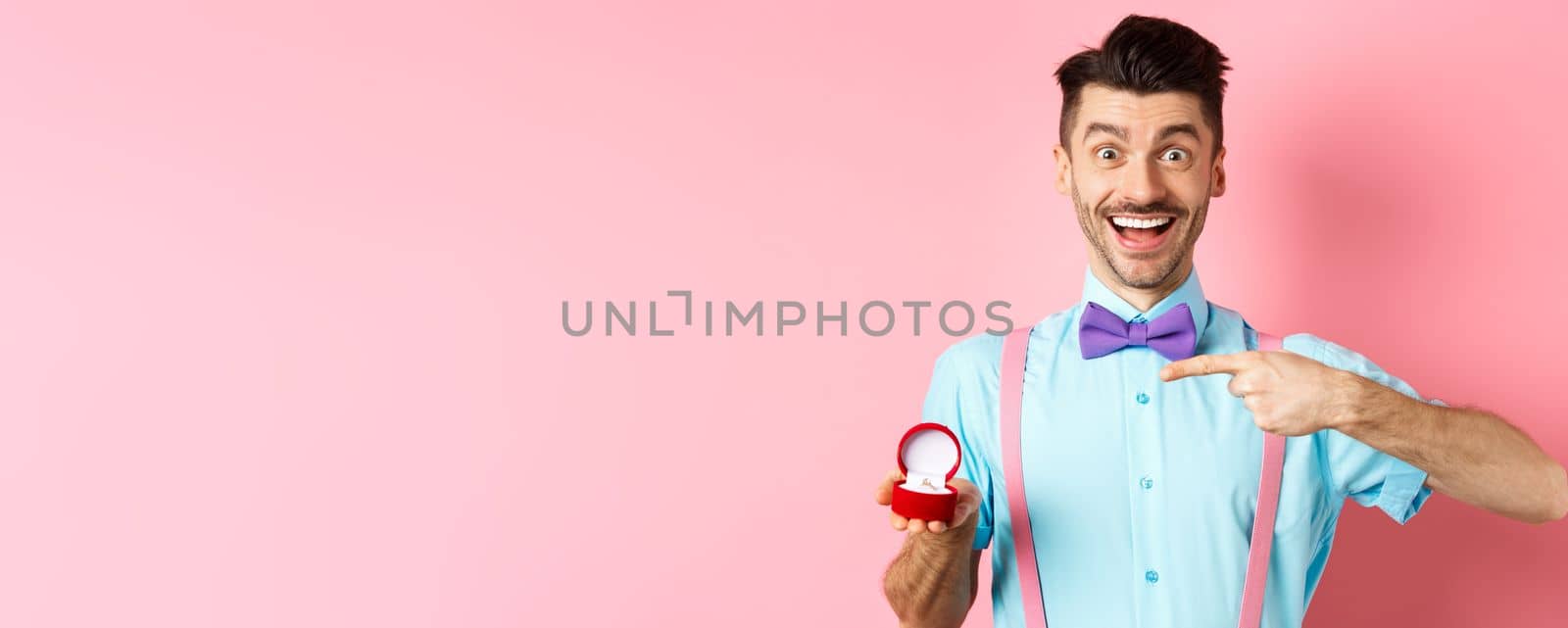 Valentines day. Cheerful young man going to make proposal, pointing finger at engagement ring and smiling excited, standing on romantic pink background by Benzoix